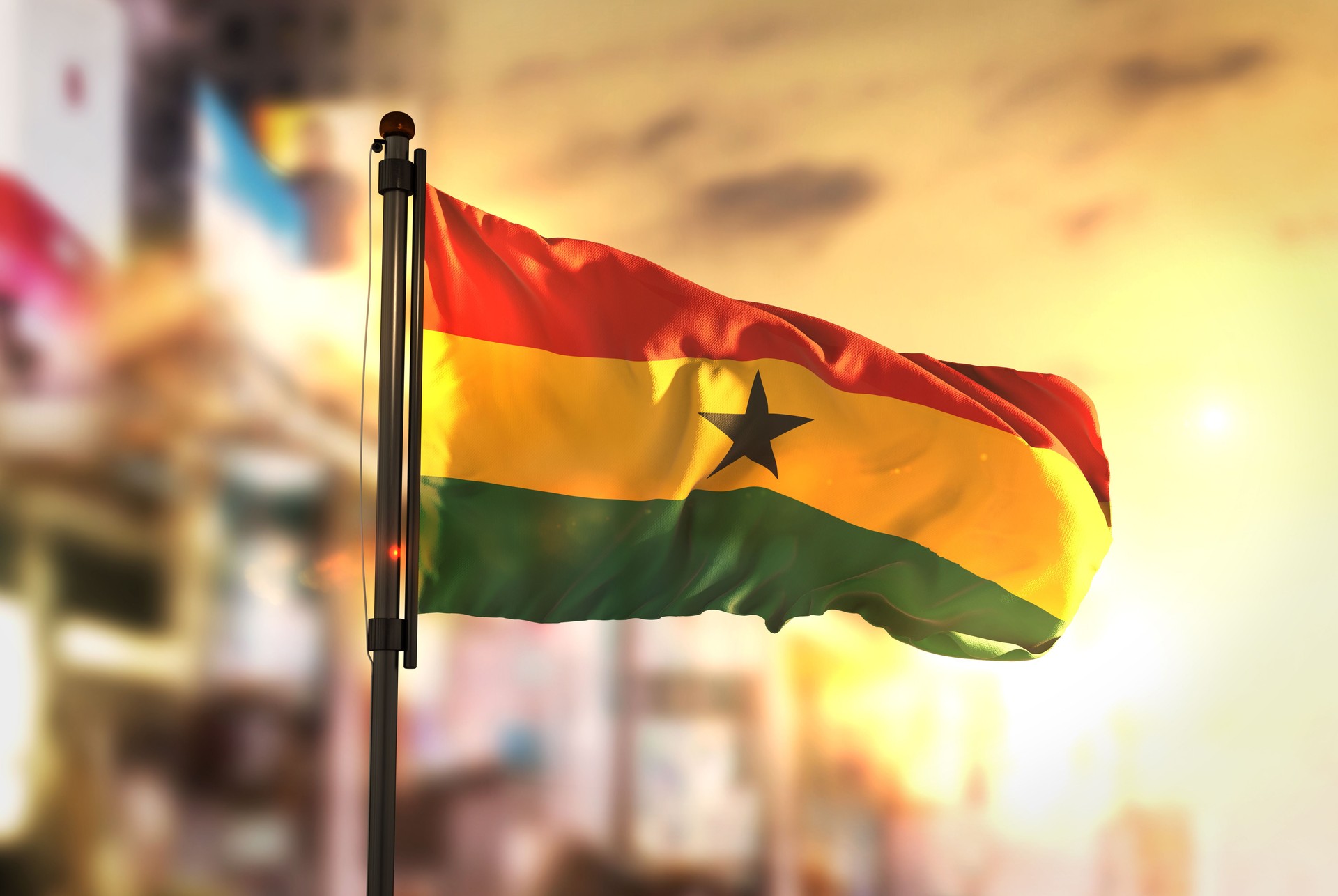 Ghana Flag Against City Blurred Background At Sunrise Backlight